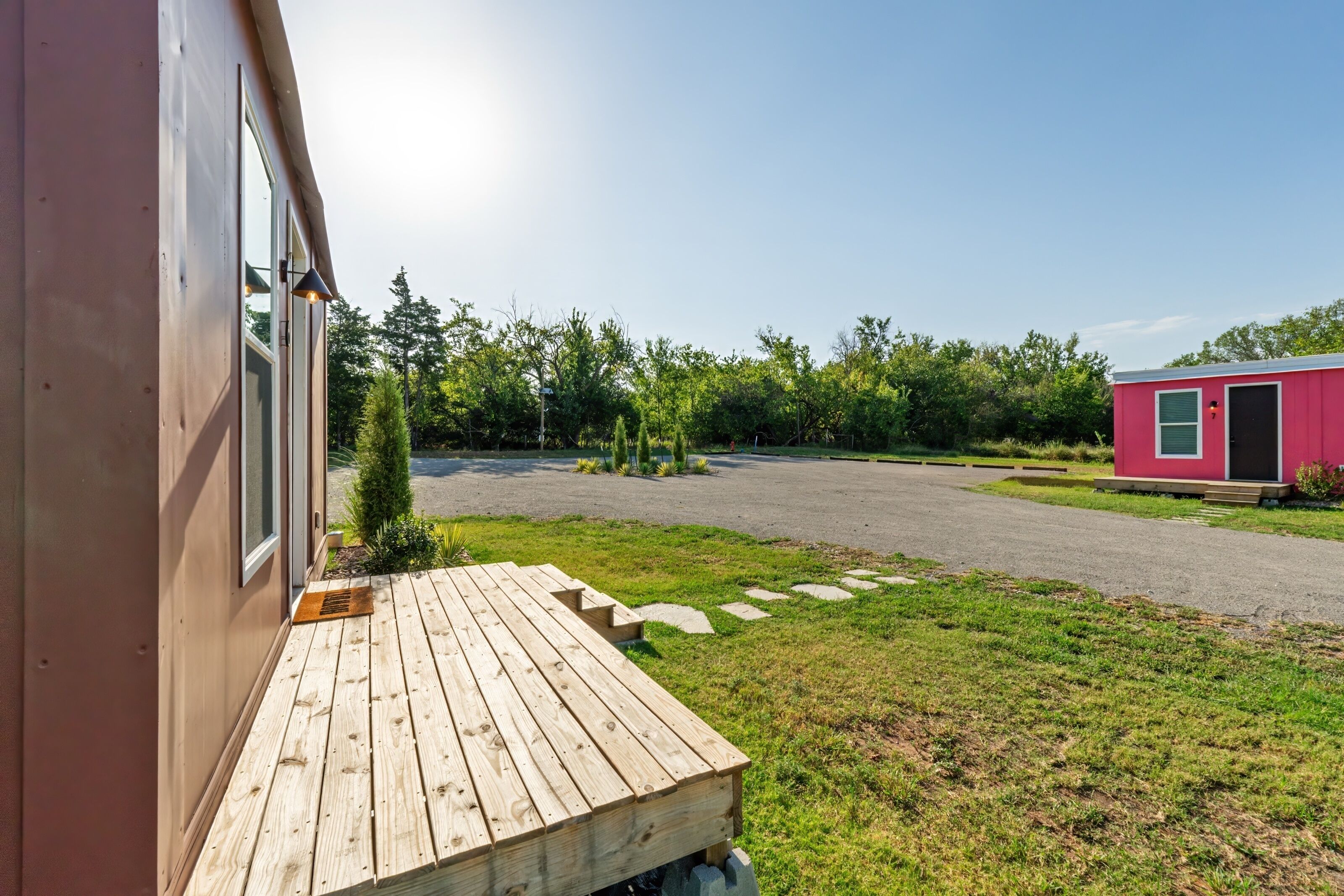 Un patio in legno che conduce a una casa con facciata rossa, un vialetto lastricato e un piccolo edificio rosa sullo sfondo.