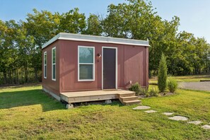Eine kleine, eingeschossige Holzhütte mit Veranda und Tür, umgeben von Wiese und Bäumen.