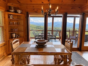 A wooden interior with a dining table, chairs, a chandelier, and a view of mountains through large windows.