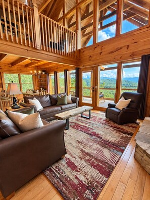 A spacious living area with a high ceiling, wooden beams, and large windows offering a view of the outdoors.