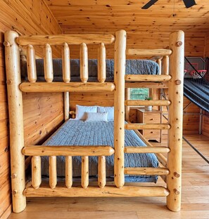 A bunk bed with a wooden ladder structure, a single bed with a quilt, and a small table with a plant.