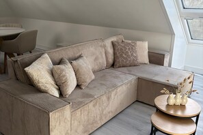 A sectional sofa with multiple cushions, a small round table with vases, and a view of the outdoors through a window.