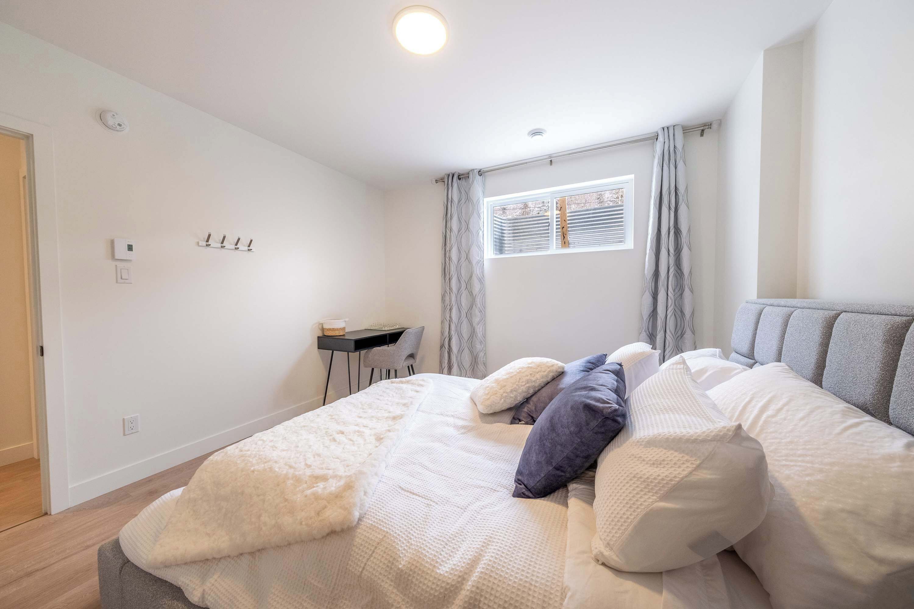 Un dormitorio con cama, almohadas, escritorio y ventana con cortinas.