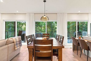 A dining area with a wooden table, chairs, and a sofa. There are large windows with curtains, and a hanging light fixture above the table.