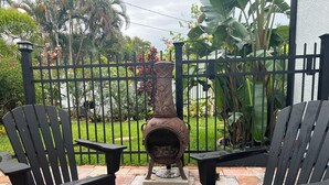 Two black Adirondack chairs on a patio with a decorative metal stove, surrounded by a black metal fence and lush greenery. - Luxurious upscale guest suite with private entrance and private tropical garden. (Gulfport)