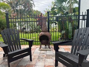 Two black Adirondack chairs on a patio with a decorative metal stove, surrounded by a black metal fence and lush greenery. - Luxurious upscale guest suite with private entrance and private tropical garden. (Gulfport)