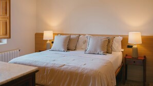 A neatly made bed with white linens and multiple pillows, flanked by two bedside tables with lamps, in a simple room with a wooden headboard and a radiator on the wall. - La Villa de Ca'n Beneït (IB)