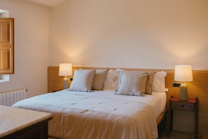 Un lit bien fait, avec du linge de lit blanc et plusieurs oreillers, flanqué de deux tables de chevet sur lesquelles sont posées des lampes, dans une chambre simple avec une tête de lit en bois et un radiateur au mur.