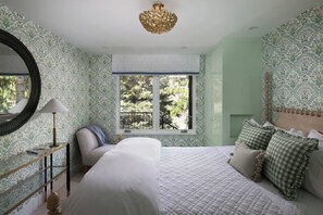 Ein Schlafzimmer mit einem großen Bett, einem Fenster mit Blick auf Bäume, einem runden Spiegel und Wandpapier mit Muster.