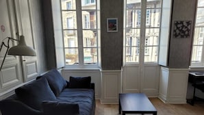 Ein Zimmer mit einem dunkelblauen Sofa, einem kleinen Tisch und einer Lampe. Fenster bieten Blick auf die Stadt.