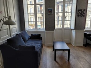 Ein Zimmer mit einem dunkelblauen Sofa, einem kleinen Tisch und einer Lampe. Fenster bieten Blick auf die Stadt.