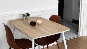 Ein Essbereich mit einem Holztisch und zwei Stühlen, eine kleine Topfpflanze an der Wand und eine durchgehende Küche hinter einer offenen Tür.