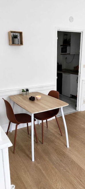 Ein Essbereich mit einem Holztisch und zwei Stühlen, eine kleine Topfpflanze an der Wand und eine durchgehende Küche hinter einer offenen Tür.