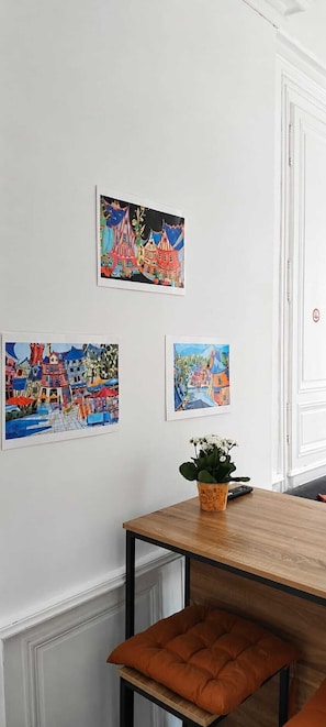 Ein Essbereich mit einem Holztisch und einer Bank, drei farbenfrohe Bilder an der Wand und ein Blumentopf auf dem Tisch.