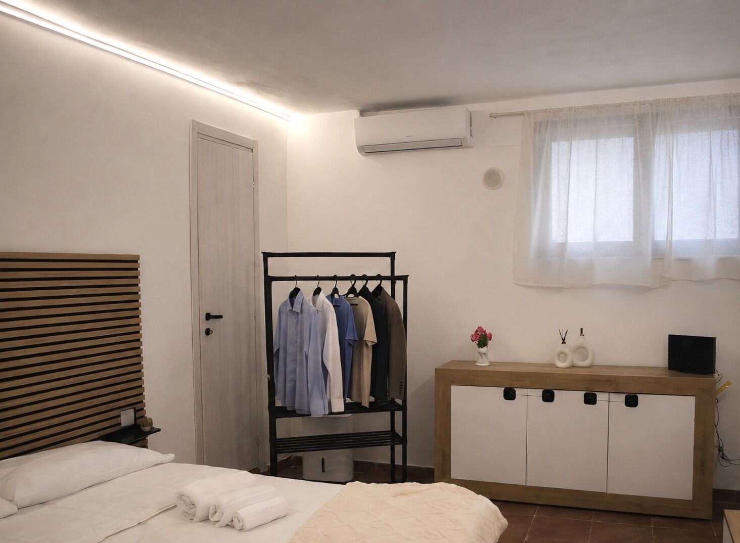 Une chambre à coucher comprenant un lit, une armoire, un portant à vêtements et une fenêtre avec des rideaux légers.