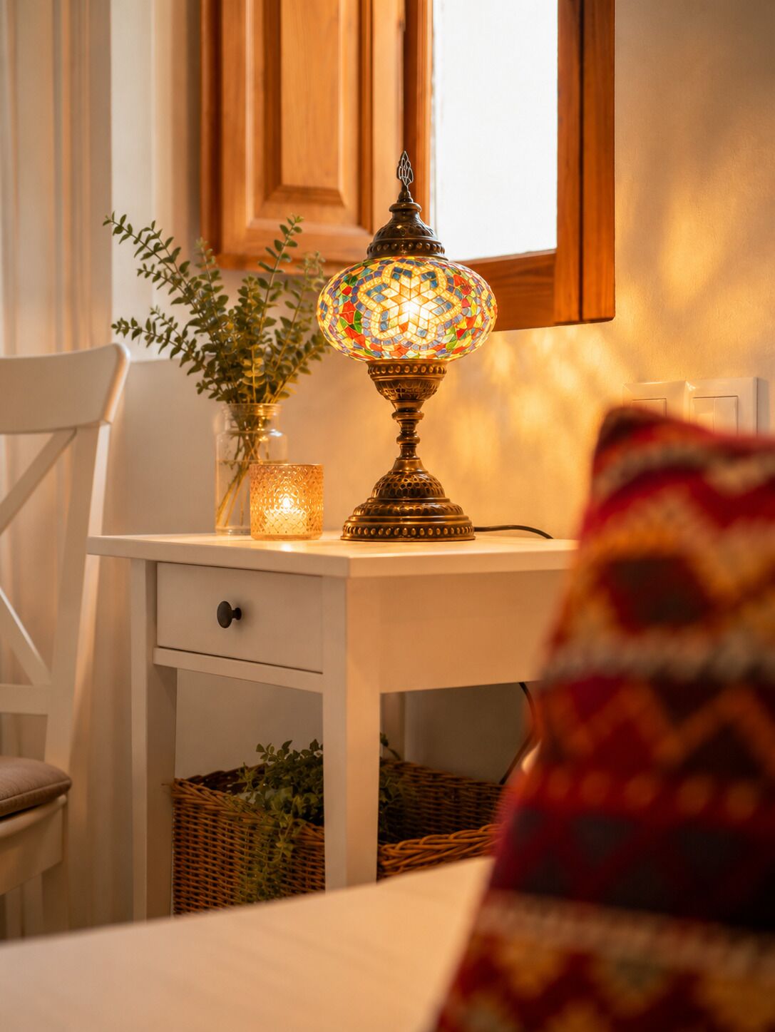 A room with a white desk, a decorative lamp, a vase with green plants, and a window with wooden shutters.