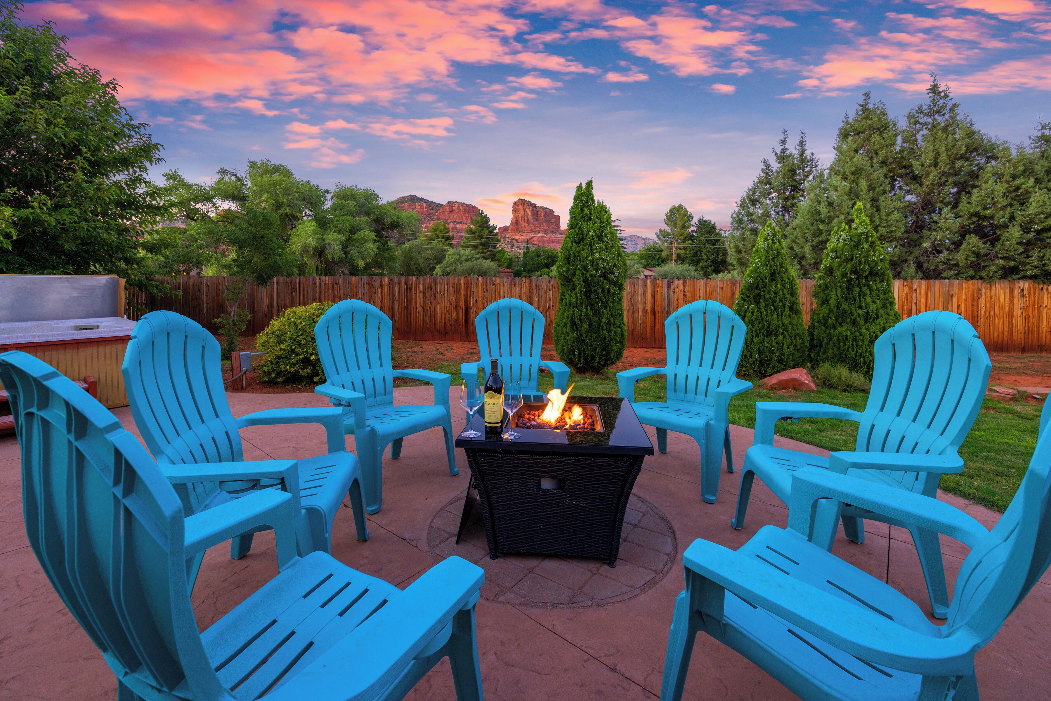 Eine Terrasse mit vier blauen Stühlen, die um eine Feuerstelle angeordnet sind, umgeben von einem Holzzaun und Grünpflanzen.