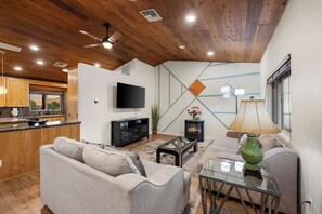 A modern living room with a sofa, a glass coffee table, a TV, and a fireplace. - Red Rock Retreat - Hot Tub, Fire Pit & Views (Sedona)