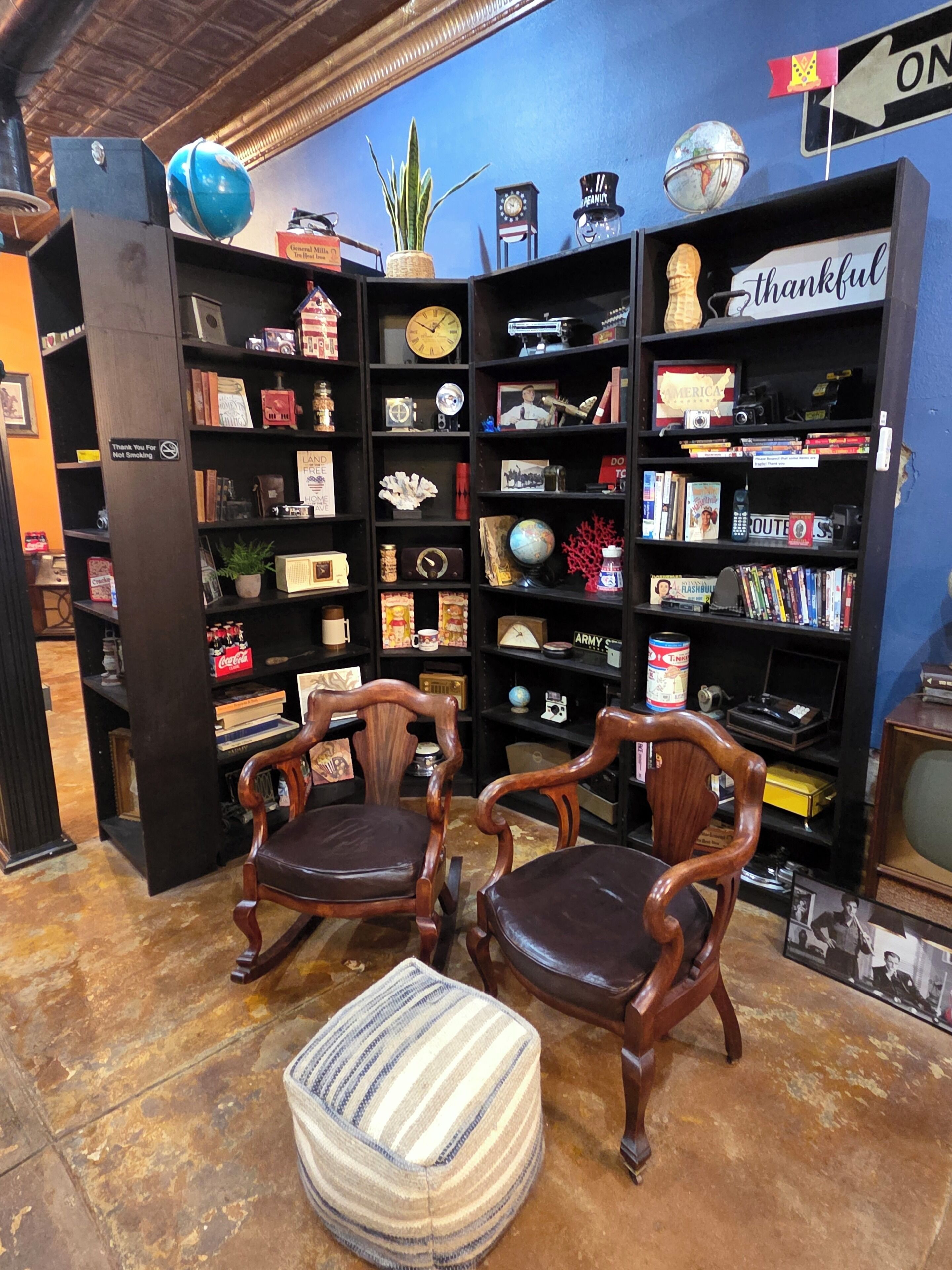 Una stanza con due sedie di legno, un poggiapiedi a righe e una libreria piena di oggetti vari.