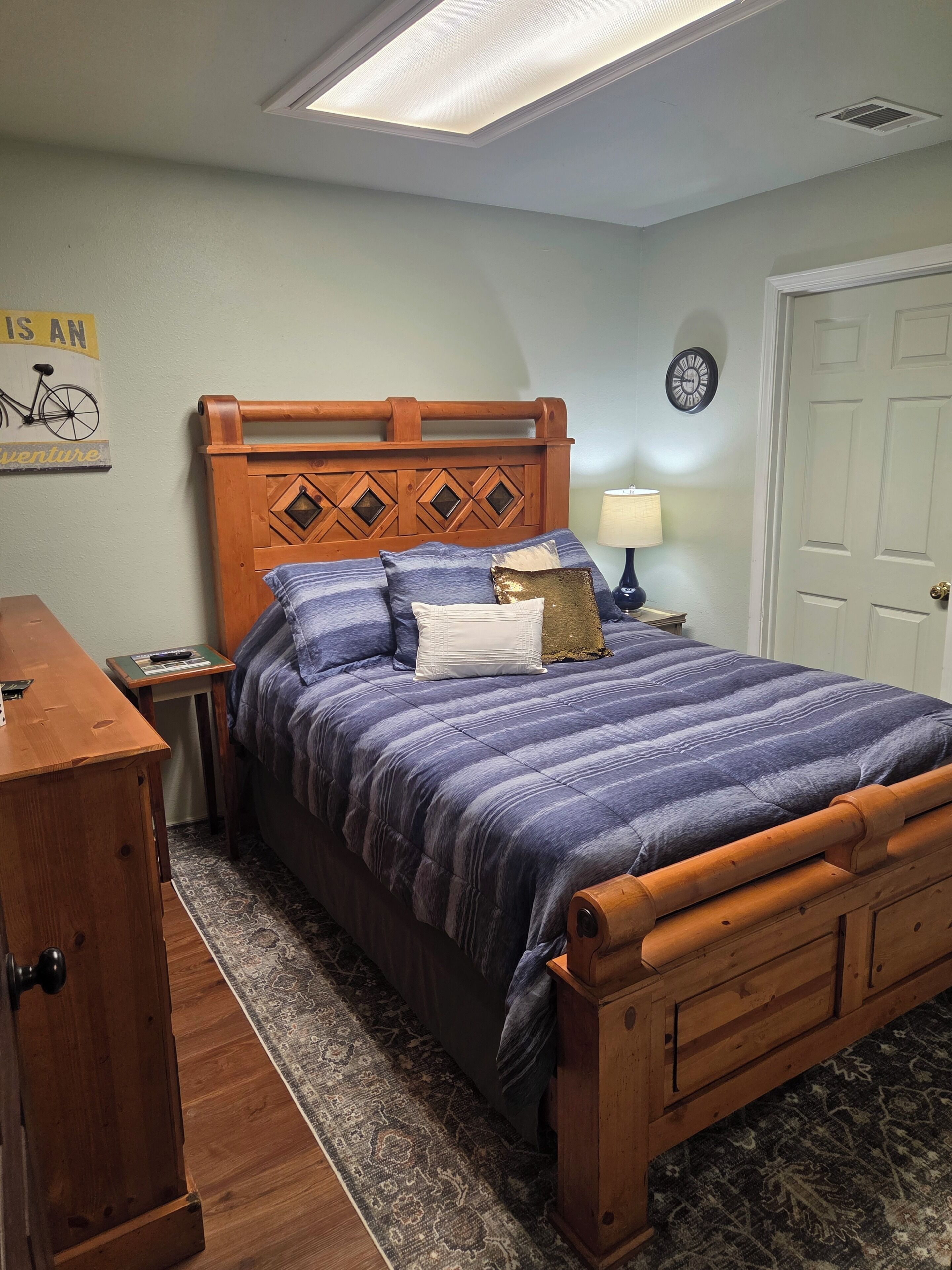 A bedroom with a wooden headboard, a bed with a blue and purple striped bedspread, a nightstand with a lamp, a wooden dresser, and a rug on the floor.