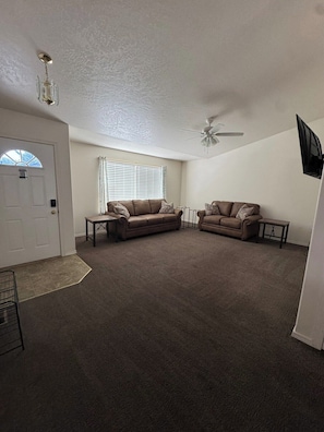 A spacious living room with two sofas, a ceiling fan, and a television mounted on the wall.