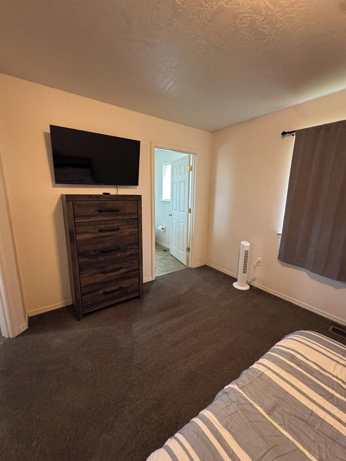 A bedroom with a bed, a dresser, a television mounted on the wall, and a door leading to another room.
