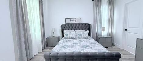 A bedroom with a tufted headboard bed, grey curtains, and a framed sign on the wall.