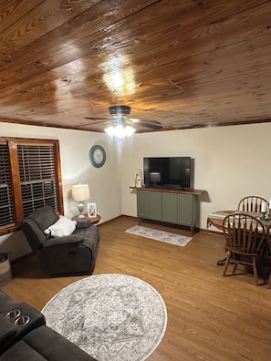 Una sala de estar con ventilador de techo, un televisor sobre un mueble bajo, una mesa de comedor con sillas y una alfombra estampada.