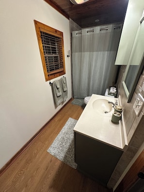 Un baño con piso de madera, un lavamanos con espejo, cortina de ducha y una ventana con persianas.