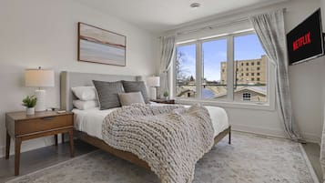 A bedroom with a bed, nightstands, a lamp, a TV, and a window with a view of buildings.