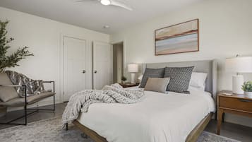 Un dormitorio con cama, una silla, una mesita de noche, una lámpara, un ventilador de techo y un cuadro en la pared.