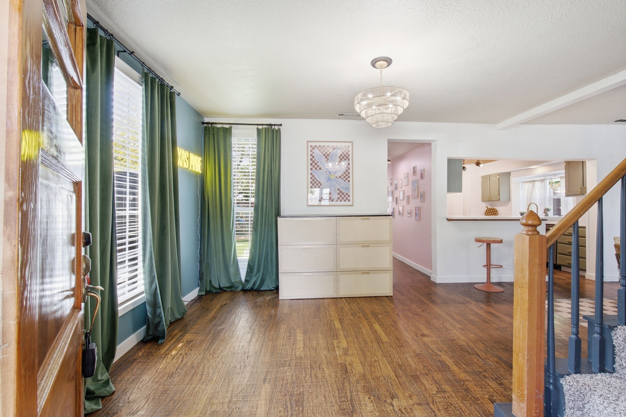 A spacious room with hardwood flooring, a chandelier, a staircase, and a kitchen area visible through an open door.