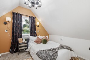 A bedroom with a sloped ceiling, a bed with pillows, a bedside table with a lamp, and a window with curtains.