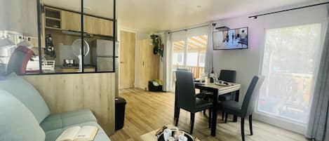 Uma sala de estar moderna com um sofá, uma mesa de jantar com cadeiras, uma TV fixada na parede e uma área de cozinha visível através de uma divisória de vidro.