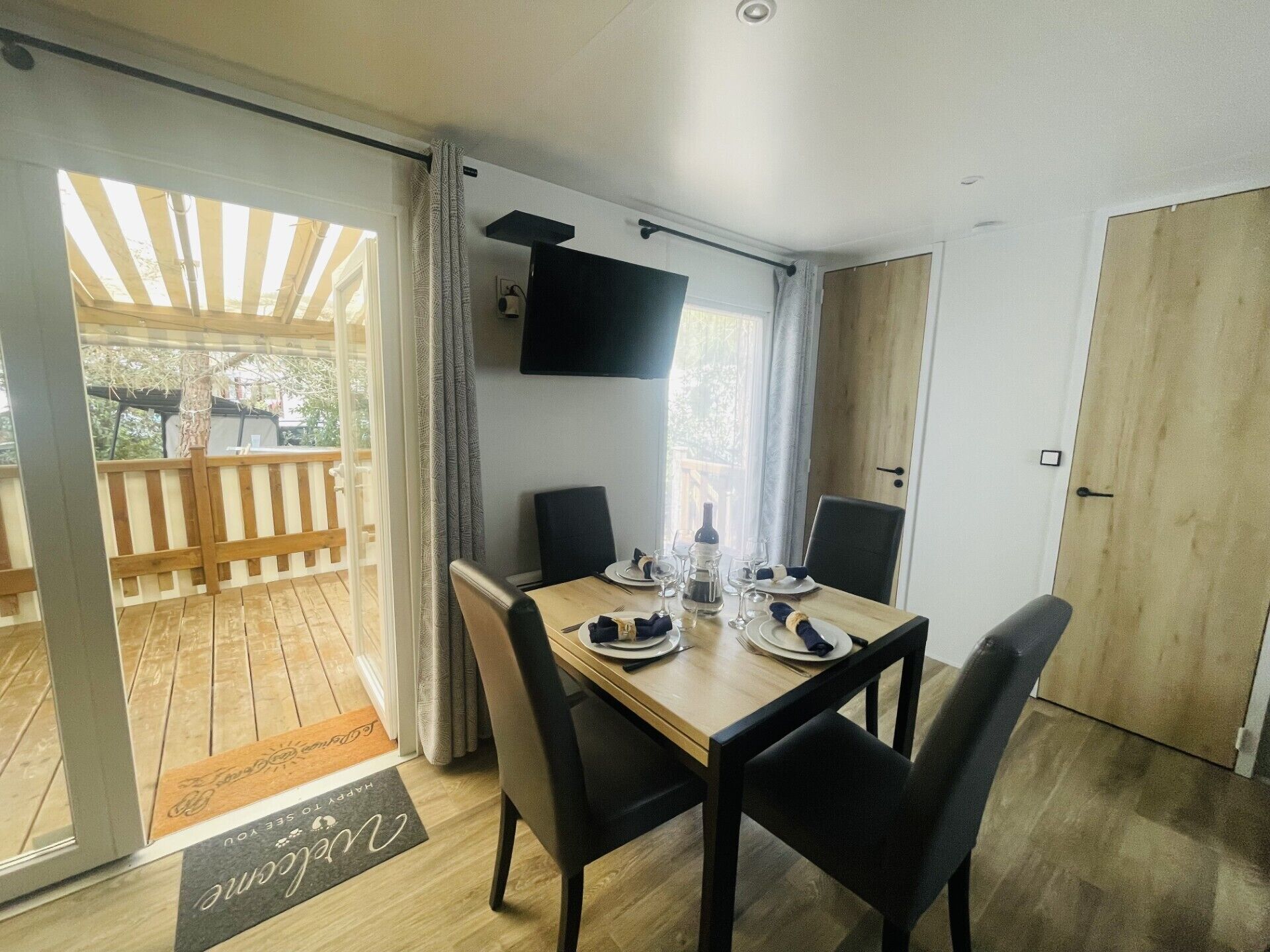 A dining area with a table set for two, a television mounted on the wall, and a sliding glass door leading to an outdoor deck.