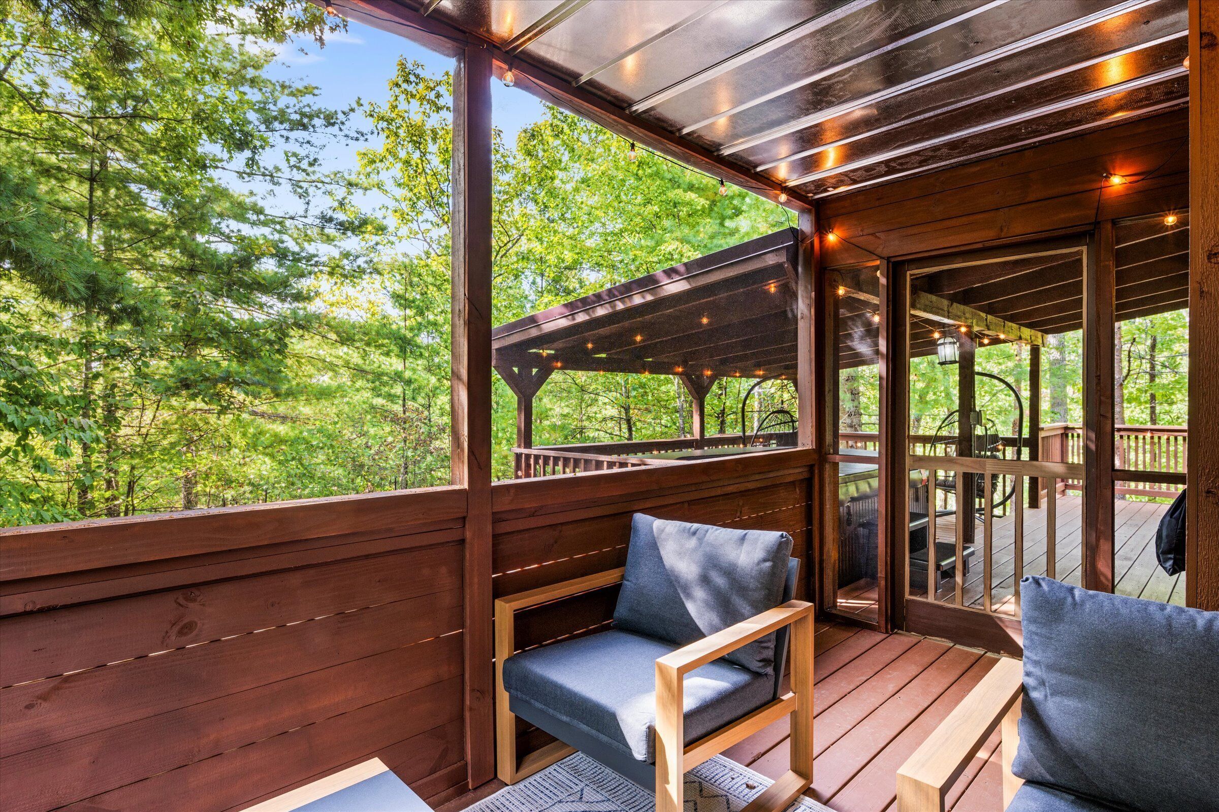 Una terraza de madera con cerramiento de vidrio, mobiliario exterior y vistas a un bosque.