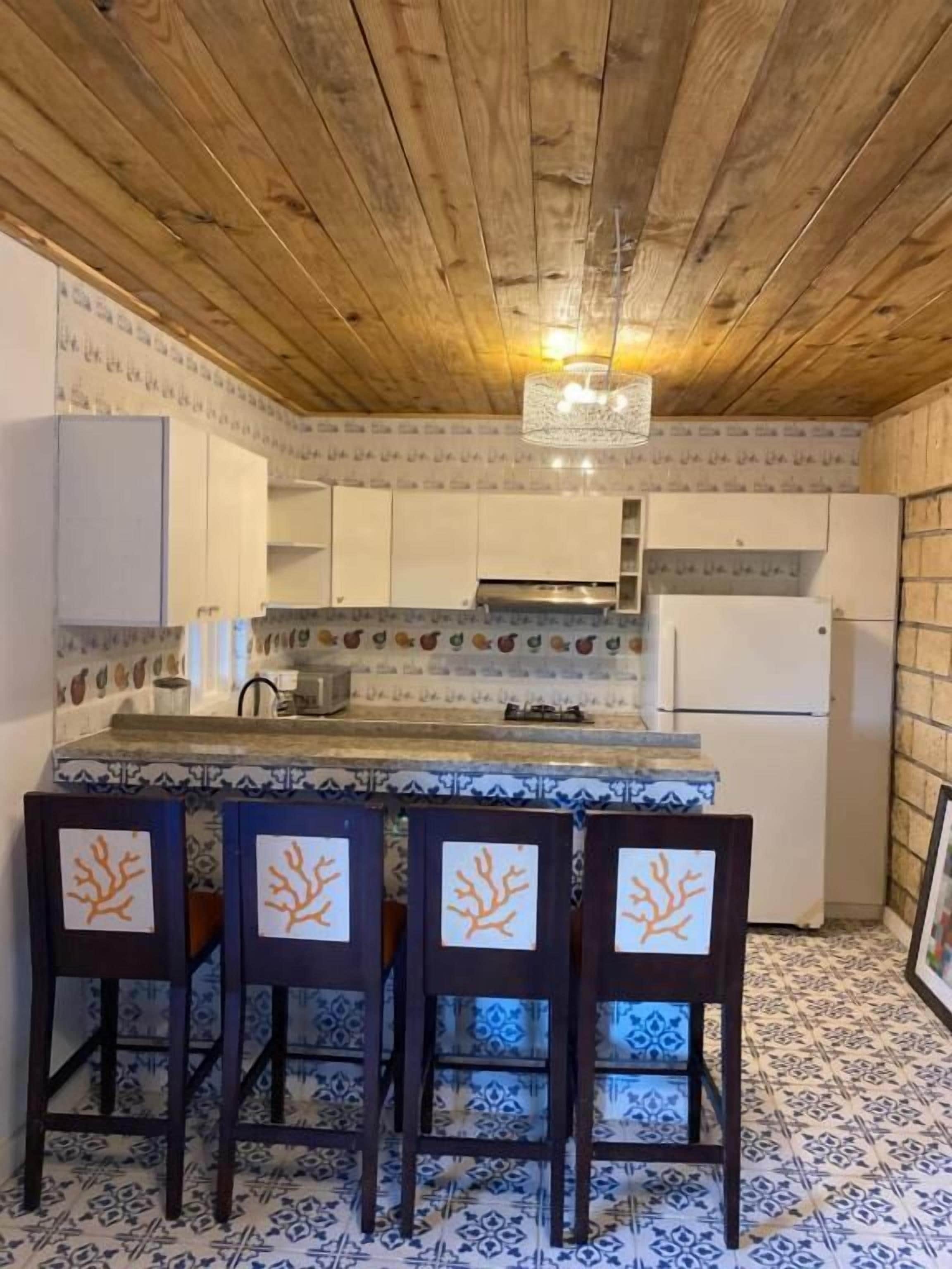 A kitchen with a wooden ceiling, white cabinets, a tiled backsplash, and a bar area with chairs.