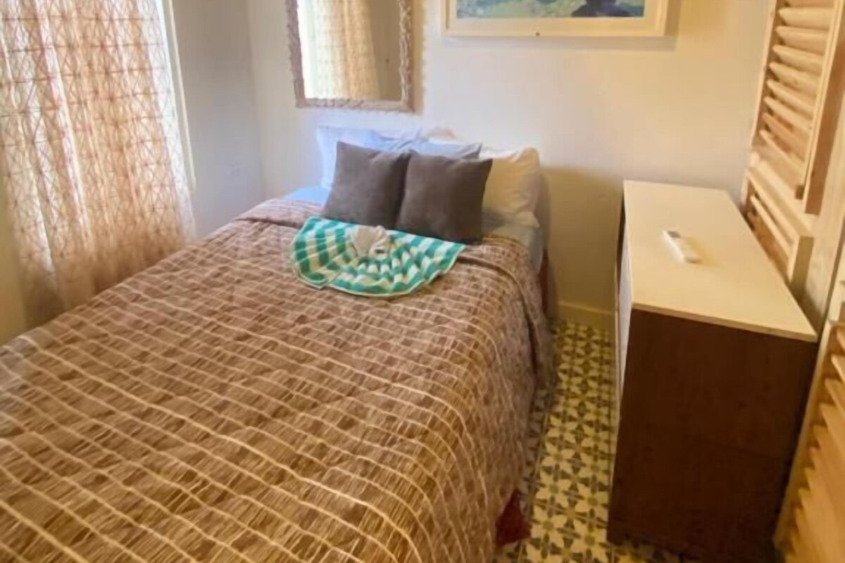 A bedroom with a bed, a bedside table, a window with curtains, and a painting on the wall.