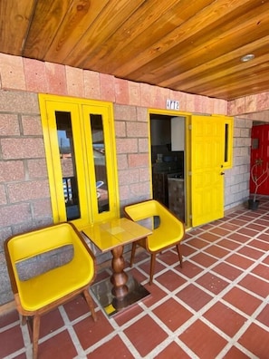 A covered outdoor seating area with yellow chairs and a table, a brick wall, and a red door. - Arco Iris Casa de Campo  (Santa Cruz)