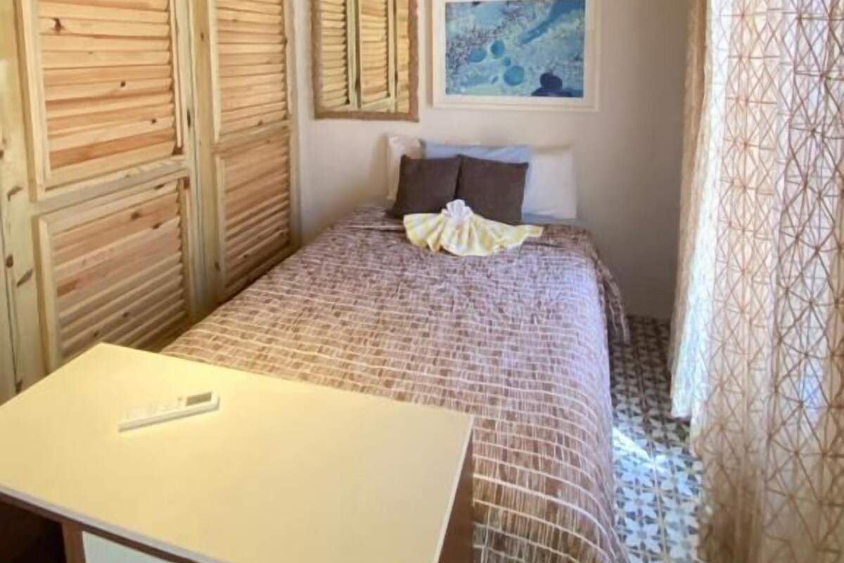 A bedroom with a wooden ceiling, a bed with a patterned bedspread, a desk, and a window with curtains.