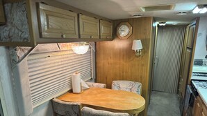 Ein kompaktes Wohnmobil-Interieur mit einem Holztisch, zwei gemusterten Stühlen, einer kleinen Küchenzeile und einer Wanduhr.