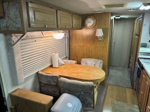 A compact RV interior with a wooden table, two patterned chairs, a small kitchenette, and a clock on the wall.