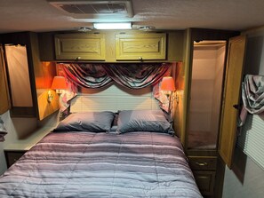 A bedroom with a bed, two bedside lamps, a window with curtains, and wooden cabinets above the bed.