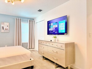 Ein Schlafzimmer mit einem Bett, einer Kommode, einem flachbildfernseher an der Wand und einem Fenster mit Vorhängen.