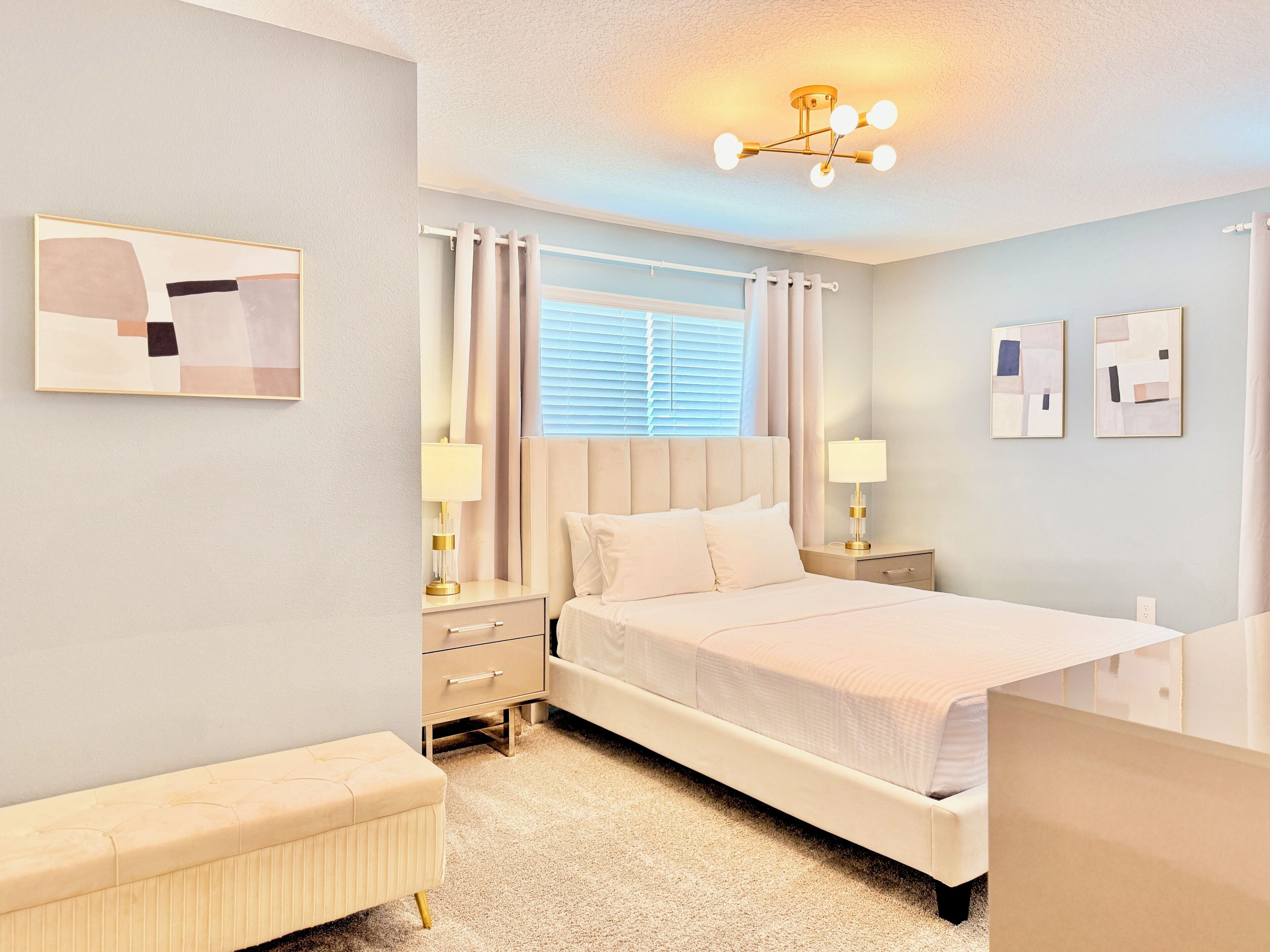 A hotel room with a bed, bedside tables, a bench, a window with curtains, and artwork on the walls.