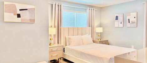 A hotel room with a bed, bedside tables, a bench, a window with curtains, and artwork on the walls.