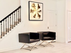 Un escalier avec une rampe noire et des murs blancs, menant à une pièce où se trouvent deux chaises noires et un tableau abstrait encadré.