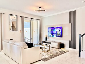 A modern living room with a white sofa, a coffee table, and a flat-screen TV mounted on the wall. - 5-bedroom townhome with Club Access, PVT Pool, BBQ Grill just 15 min from Disney (Kissimmee)