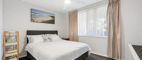 Una camera da letto con un letto, un comodino, una finestra con tende, una ventola a soffitto e un quadro appeso al muro.