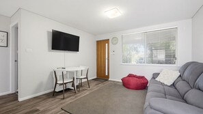 Een woonkamer met een grijze bank, een rode pluche stoel, een witte eettafel met stoelen, een flatscreen televisie aan de muur en een raam met jaloezieën.